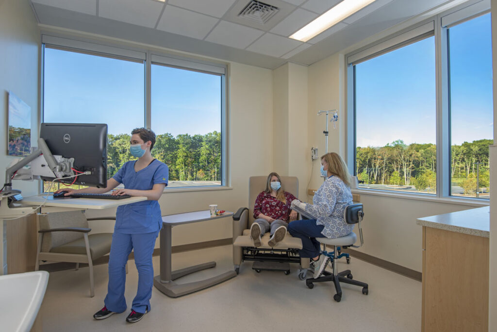 A technician works on their computer while a nurse tends to a patient receiving IV treatment inside of a medical infusion bay