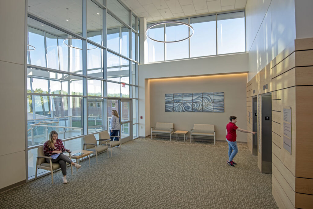 Patient waiting area inside of a medical office building. A member of hospital staff exits to the vestibule (which is seen through floor-to-ceiling windows) while one patient is seated reading a magazine, and another patient presses a button to call an elevator.