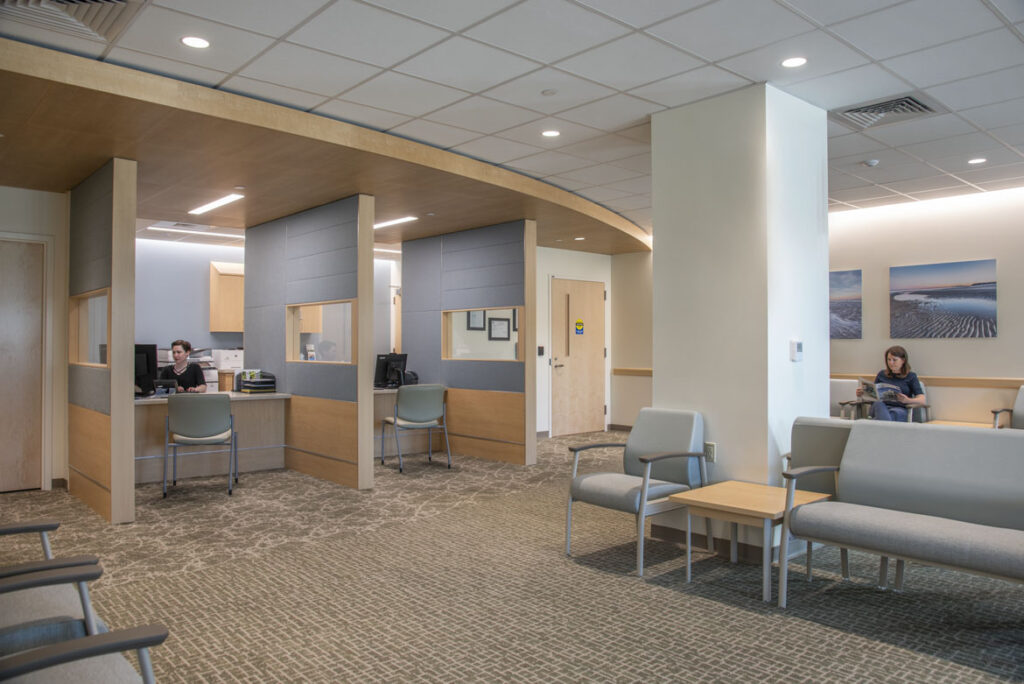 A patient registration desk within a medical office building. A receptionist works at their desk while a patient reads a magazine while seated.