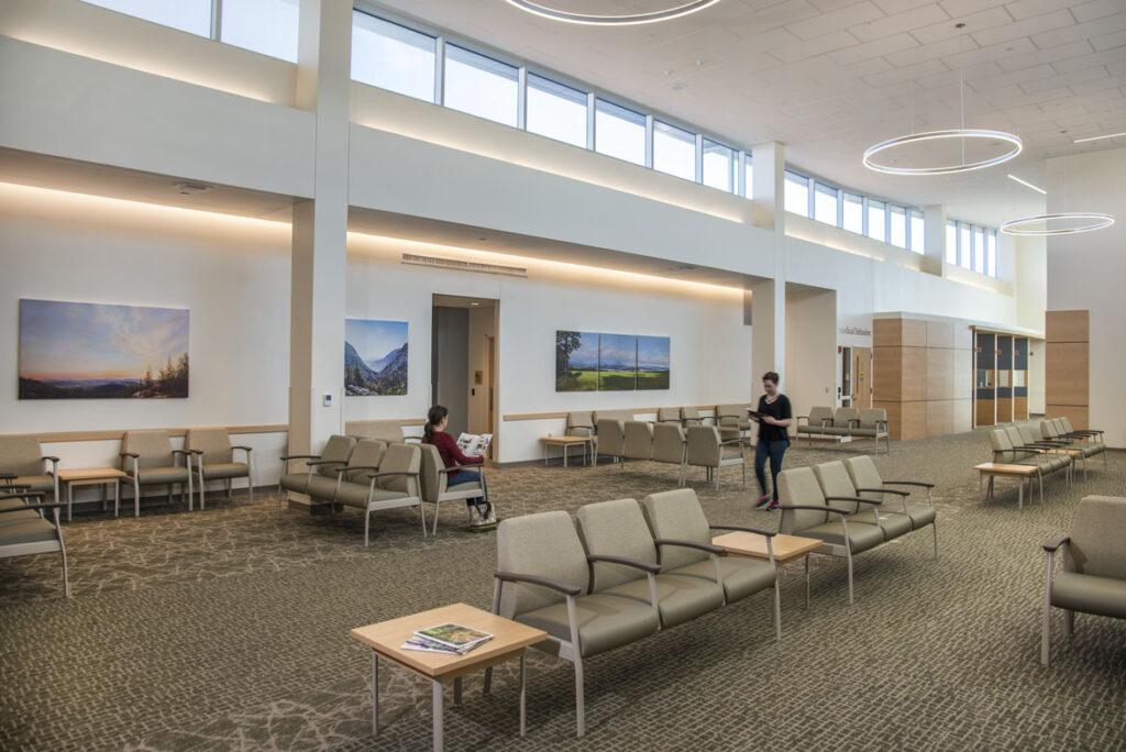 Open office waiting area with seating. One patient sits and reads a magazine while another walks around holding a tablet computer.